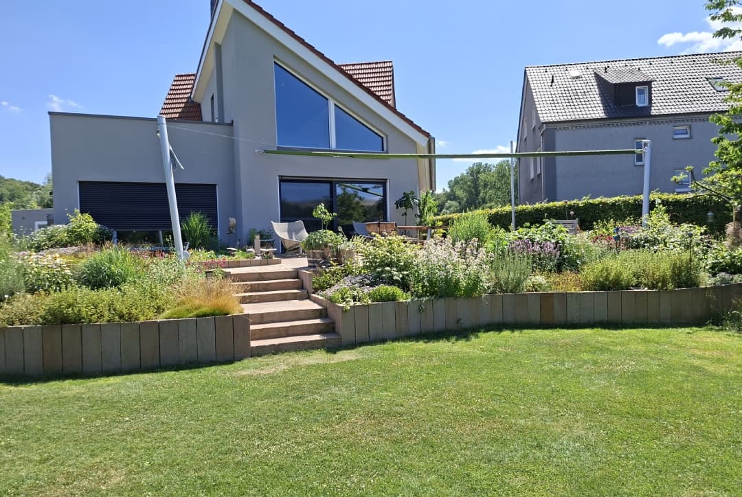 Gartenanlage mit Natursteintreppe, Holz-Palisaden-Beeteinfassung und Rasenfläche vor Einfamilienhaus
