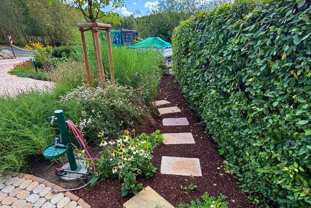 Trittplatten-Weg entlang einer Hecke mit Wasserzapfstelle und Staudenbeet